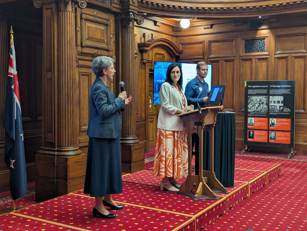Dr Black stands in profile, speaking into a microphone. Dr Parmar and Dr Cahill stand in profile next to her and behind a podium. All stand on red carpet with ornate wood paneling and an NZ flag behind them.